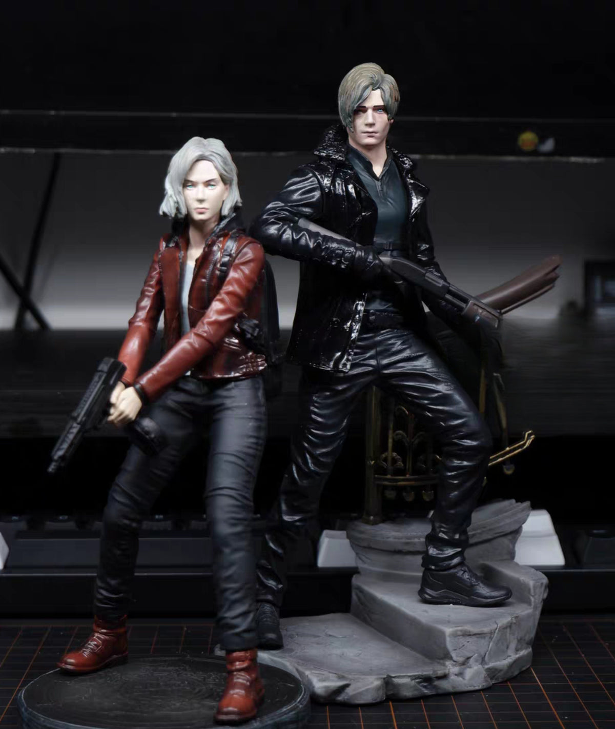 Leon Kennedy Grace Ashcroft Resident Evil action figures posed together on a display base against a dark studio background. The figure on the left has silver hair and wears a red jacket with black pants and boots, holding a gun.
