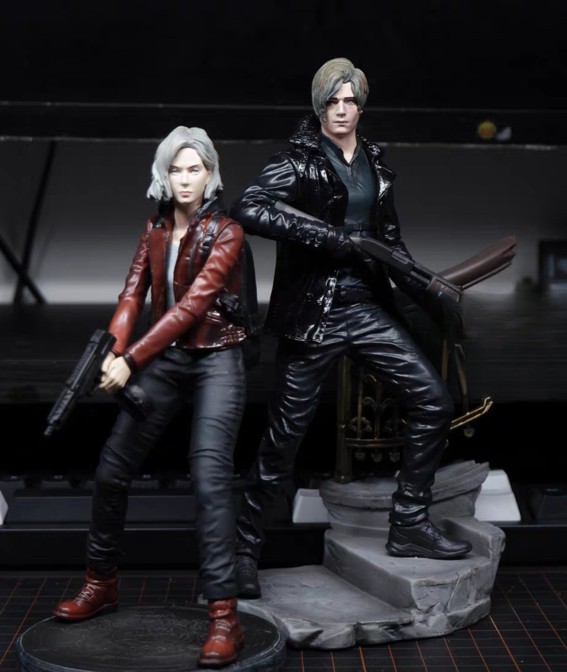 Leon Kennedy Grace Ashcroft Resident Evil action figures posed together on a display base against a dark studio background. The figure on the left has silver hair and wears a red jacket with black pants and boots, holding a gun.