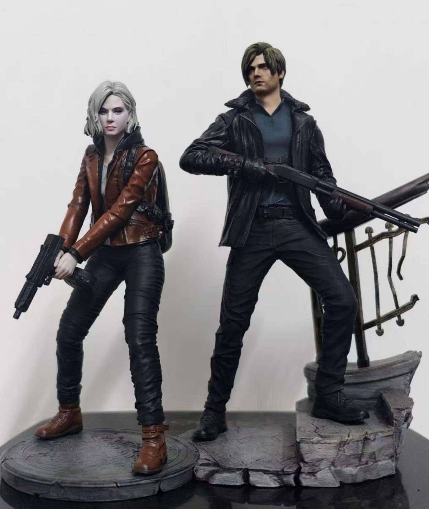Leon Kennedy Grace Ashcrof action figures posed on a display base against a plain light background. The figure on the left has pale skin, short gray hair, and wears a brown leather jacket, black pants, and brown boots while holding a gun.