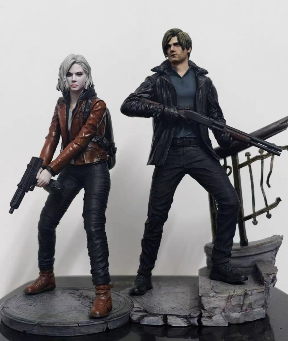 Leon Kennedy Grace Ashcrof action figures posed on a display base against a plain light background. The figure on the left has pale skin, short gray hair, and wears a brown leather jacket, black pants, and brown boots while holding a gun.