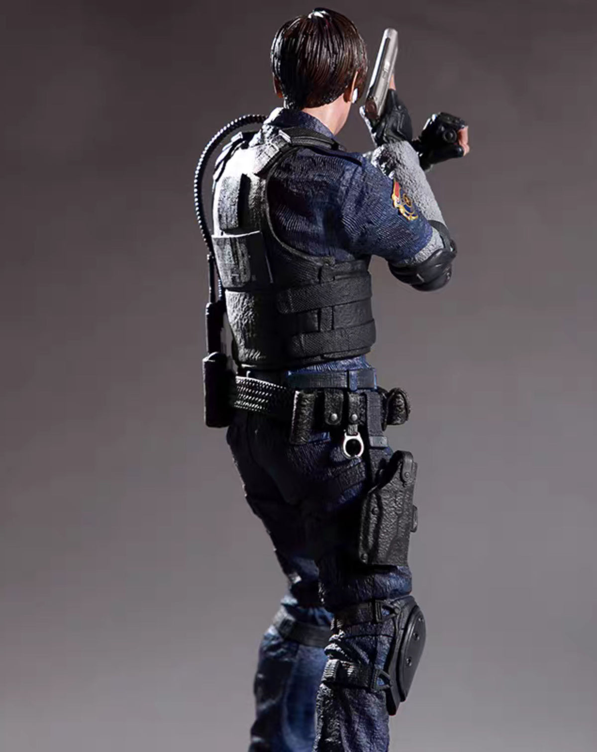 A collectible action figure oLeon Kenedy shown from the back and side, holding a silver pistol, wearing a dark blue uniform, black tactical vest, and dark pants against a gray studio background.