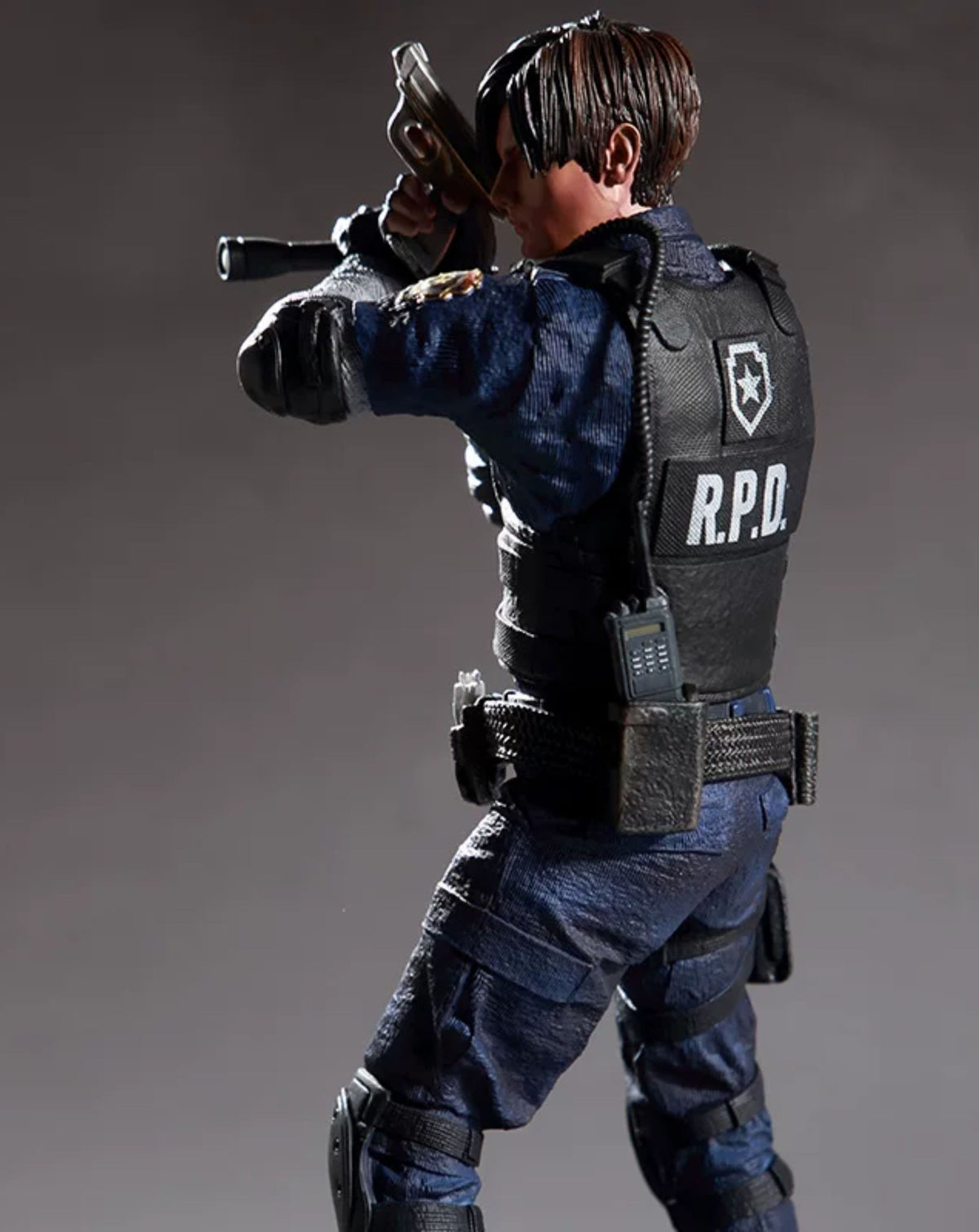 A detailed action figure of Leon Kennedy from Resident Evil, shown from a dynamic three-quarters back view. The figure is wearing a dark blue uniform, a black tactical vest with 'R.