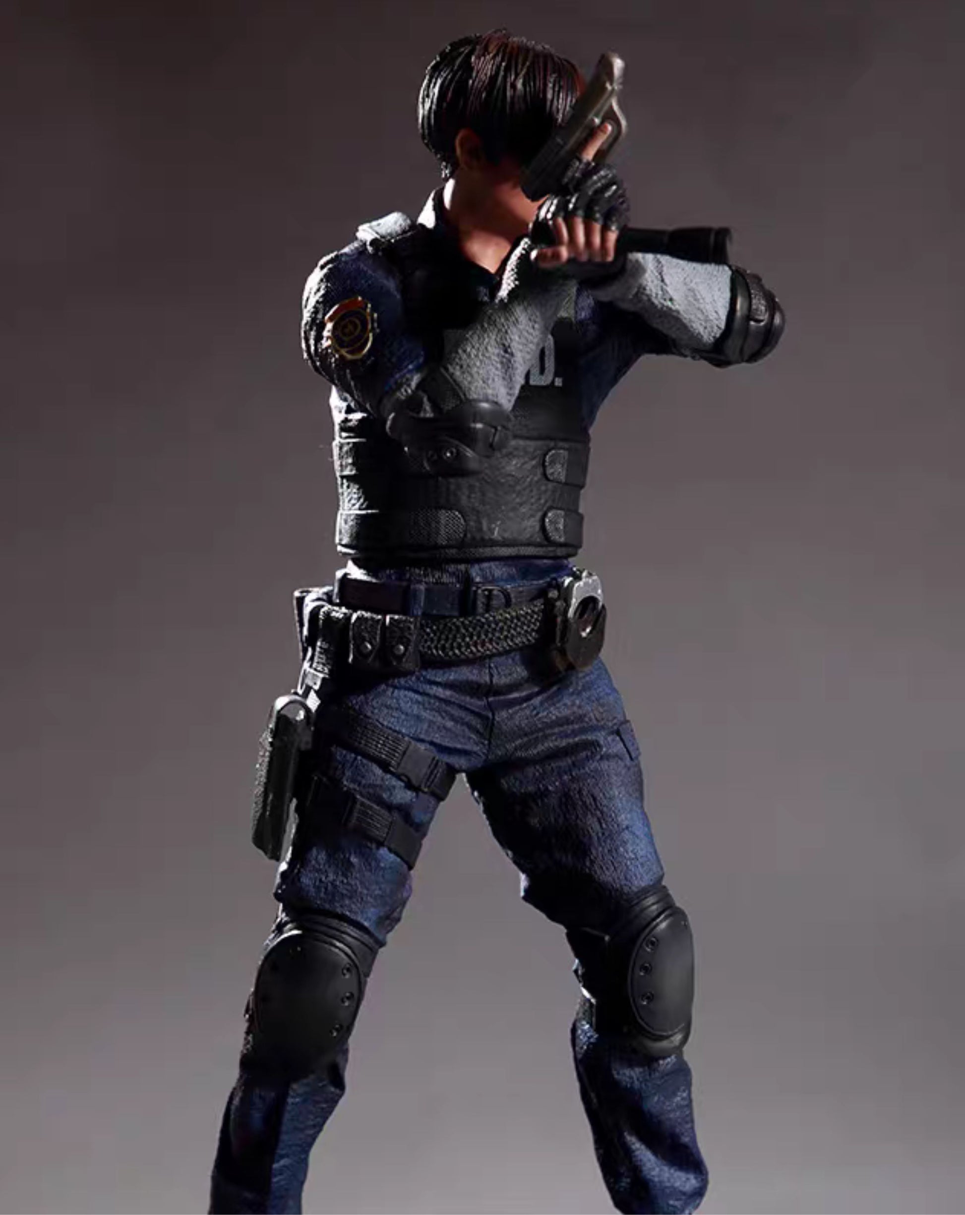 A detailed action figure of aLeon S. Kennedy from Residen Evil, in a crouched combat pose, wearing a dark gray and black vest, blue jeans, and black knee pads, holding a pistol to his face against a dark gray studio background.