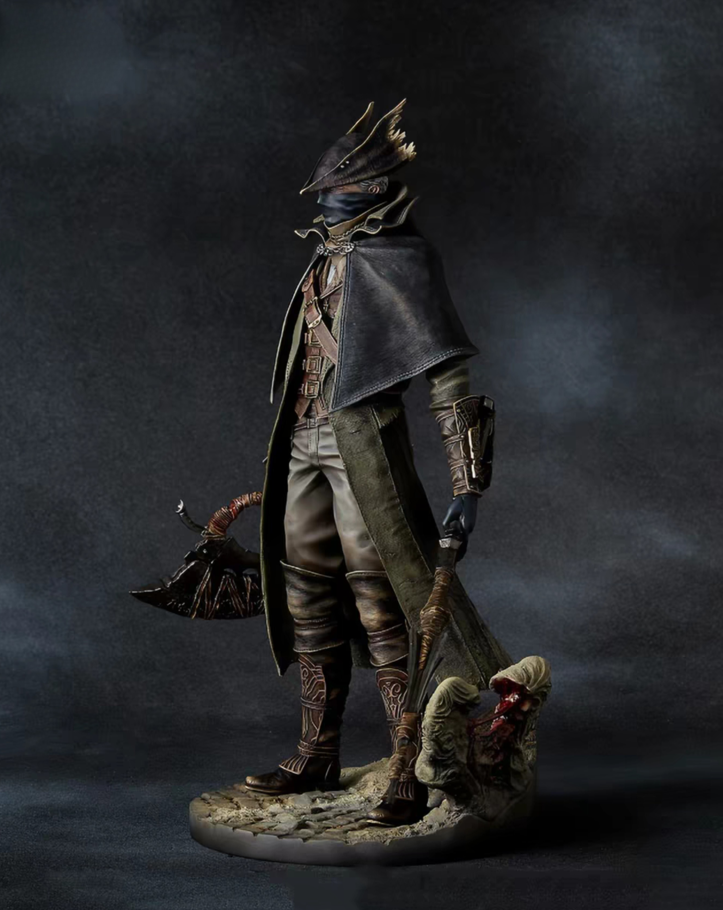 A detailed collectible figurine of a Hunter from Bloodborne dark fantasy character standing on a rocky base, holding a large axe, against a dark smoky studio background.