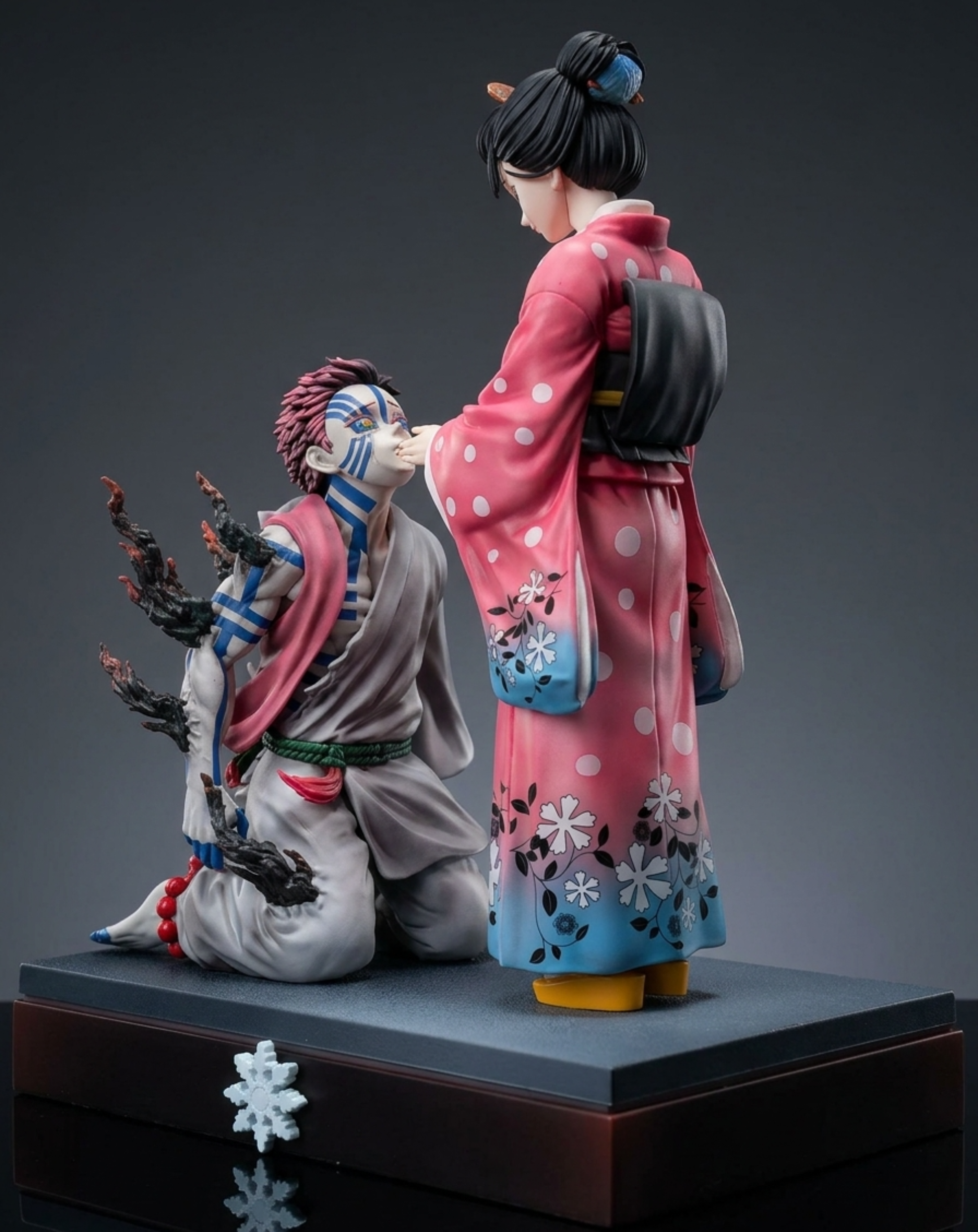 A detailed collectible figurine of 	Akaza and Koyuki from Demon Slayer, displayed on a dark base against a dark gray studio background. Akaza is kneeling in a gray robe with blue and white markings, while Koyuki stands in a pink and blue floral kimono.