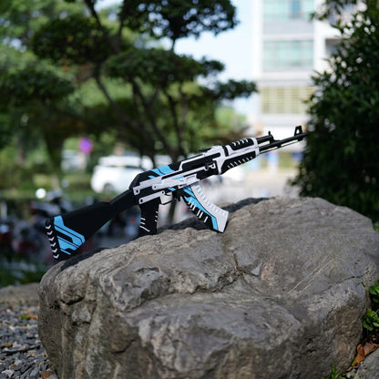 AK47 Vulcan CS2 life size replica resting on large rock outdoors — white and blue geometric skin, blurred urban street and trees background, full length side profile view, natural daylight