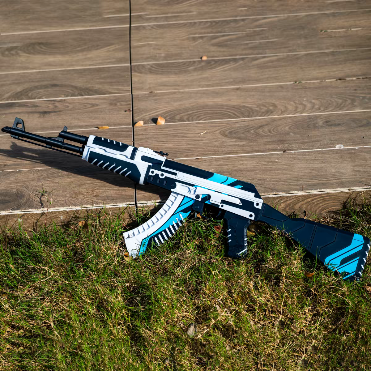 AK47 Vulcan CS2 life size replica laying on grass beside wooden deck — white and blue geometric skin detail, full length diagonal view, stock and magazine clearly visible, natural daylight