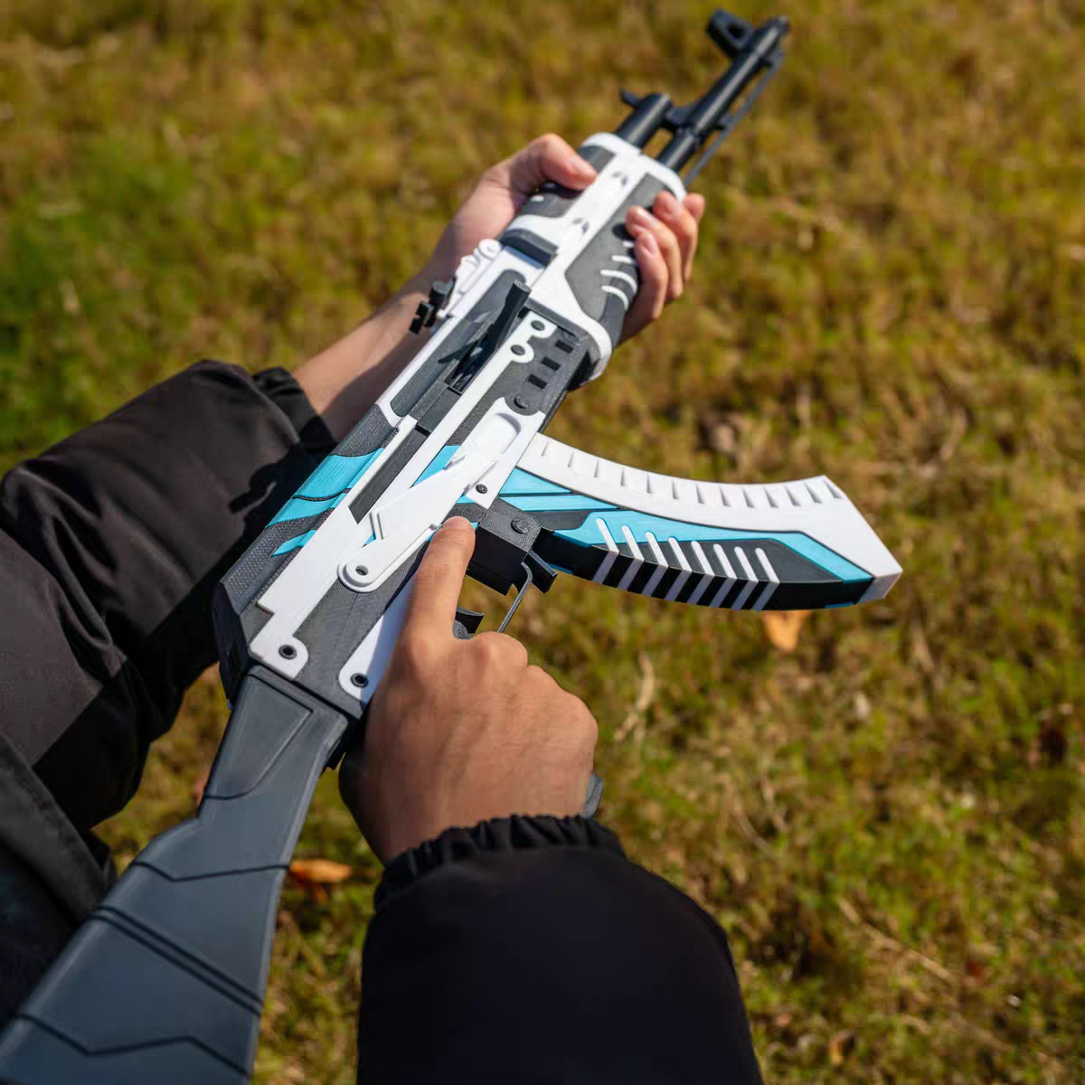 Both hands holding AK47 Vulcan CS2 life size replica from above — white and blue geometric skin detail, removable magazine visible, grass background, overhead angle showing full body of weapon